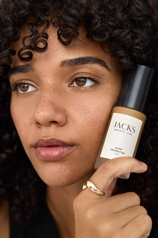 #farbwunsch_n-6, #farbwunsch_mittlerer-bis-dunkler-hauttyp, Frau trägt JACKS beauty line Glow Foundation N6 im Gesicht und Flakon in der Hand