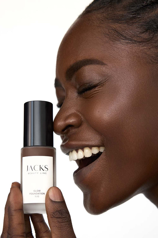 #farbwunsch_n-10, #farbwunsch_sehr-dunkler-hauttyp, Frau trägt JACKS beauty line Glow Foundation im Gesicht und Flakon in der Hand