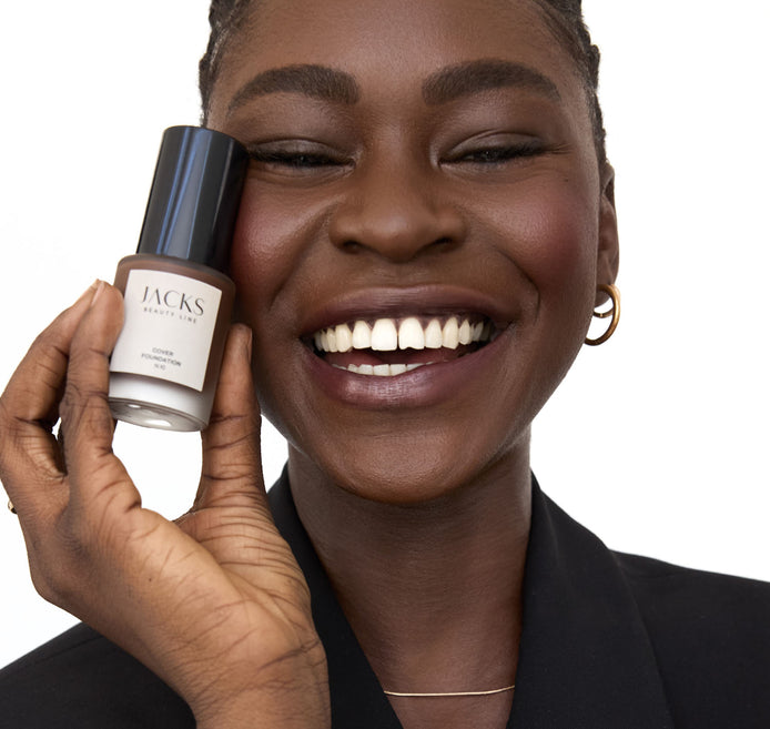 Schwarze Frau lächelt und hält die JACKS beauty line Cover Foundation neben ihr Gesicht