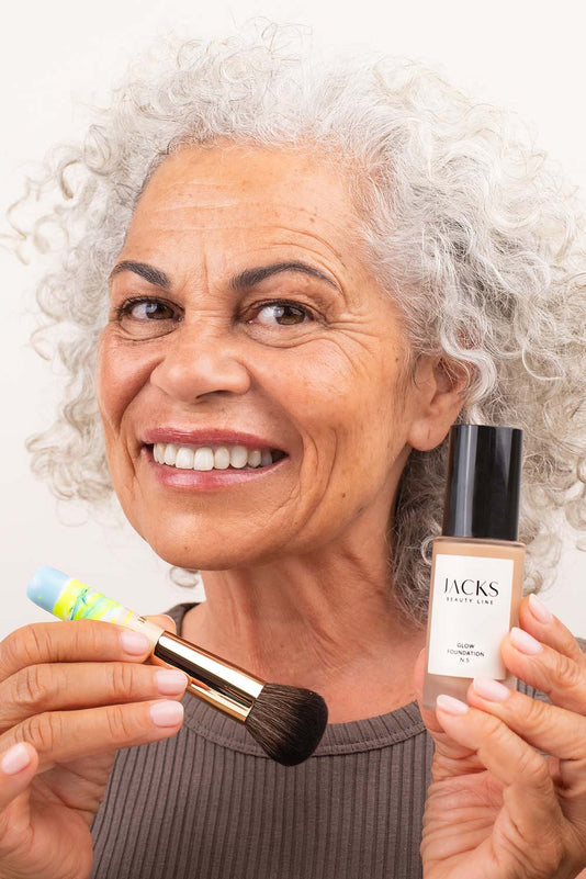 #farbwunsch_n-5, Frau Ü50 hält die JACKS beauty line Glow Foundation N.5 und den Mini Foundationpinsel