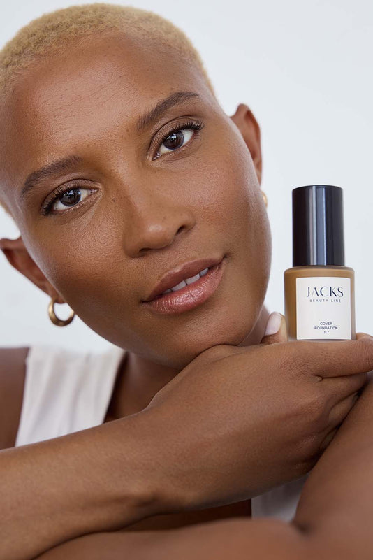 #farbwunsch_n-7, Schwarze Frau hält JACKS beauty line Cover Foundation N7 Flakon neben ihrem Gesicht