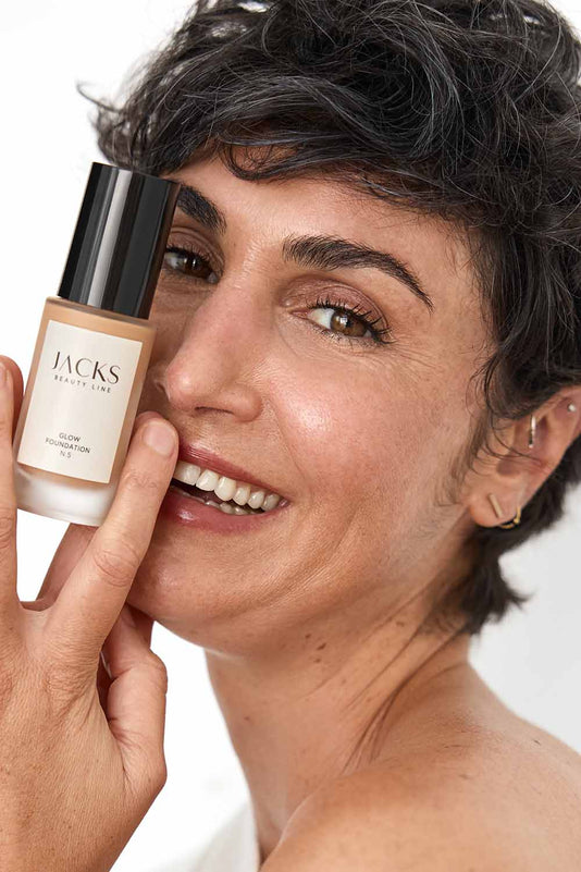 #farbwunsch_n-5, #farbwunsch_mittlerer-bis-dunkler-hauttyp, Frau trägt JACKS beauty line Glow Foundation N5 im Gesicht und Flakon in der Hand