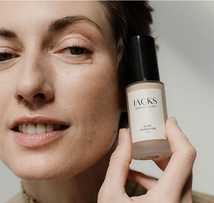 Frau hält die JACKS beauty line Glow Foundation in der Hand neben ihrem Gesicht