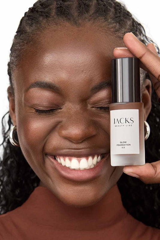 #farbwunsch_n-9, #farbwunsch_dunkler-hauttyp, #farbwunsch_sehr-dunkler-hauttyp, Frau trägt JACKS beauty line Glow Foundation im Gesicht und Flakon in der Hand 