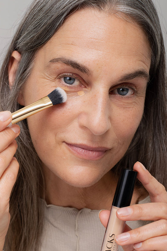 #farbwunsch_n-4, Grau-braunhaarige Frau trägt den JACKS beauty line Concealer mit dem Concealerpinsel auf