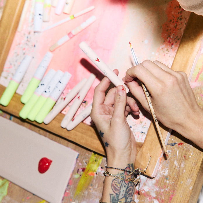 Close-up von zwei Händen, die einen Make-up Pinsel mit roten Herzen bemalt