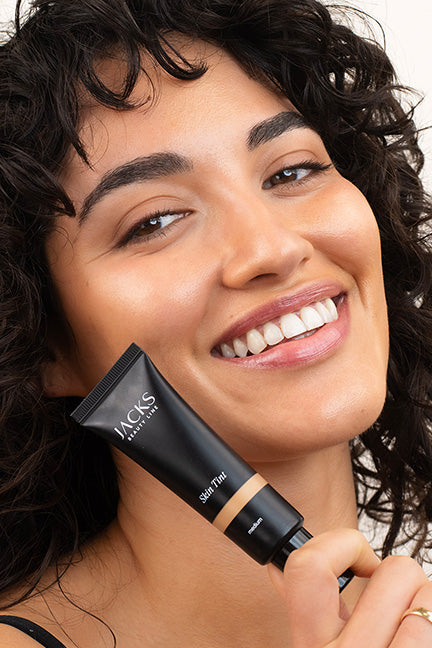 Lächelnde Frau trägt das Skin Tint in Medium auf der Haut und hält das Produkt in der Hand 